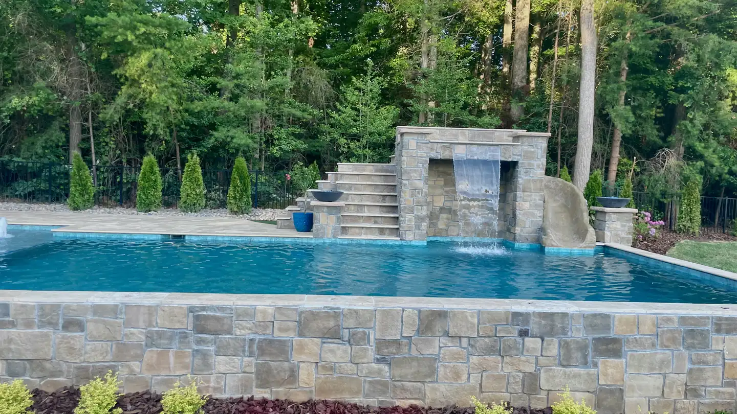 A pool with a stone waterfall wall and raised stone coping, backed by woods