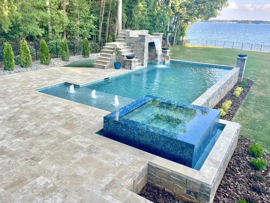 A pool with a raised tile spa and a stone waterfall on a lakefront lawn